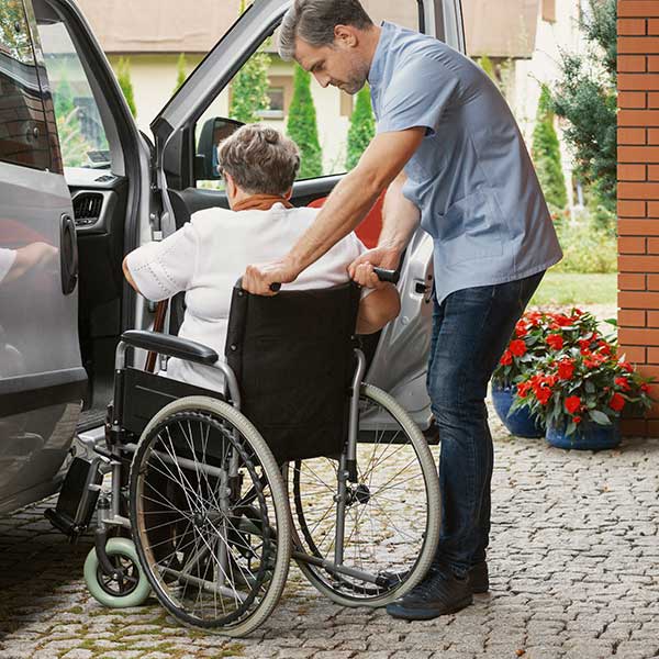 Male helping elder woman in wheel chair to car